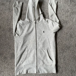 Grå hoodie med dragkedja - Snygg och bekväm grå hoodie från Polo Ralph Lauren med dragkedja och huva. Den har en liten broderad logga på bröstet och praktiska fickor framtill. Perfekt för en avslappnad stil.
