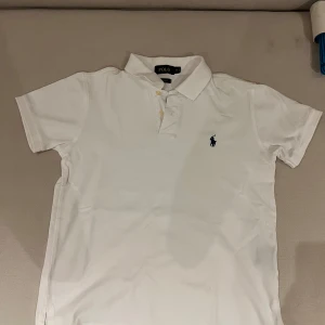 Vit kortärmad pikétröja från Polo - Säljer en klassisk vit pikétröja från Polo med korta ärmar och en broderad logga på bröstet. Tröjan har en stilren design med krage och knappar framtill. Perfekt för en avslappnad men ändå stilren look.