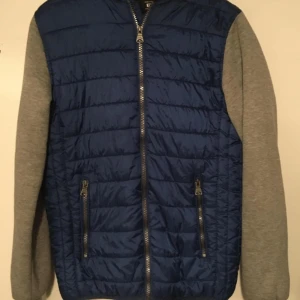 Blå quiltad jacka från FSBN - Snygg blå quiltad jacka från FSBN med gråa ärmar. Jackan har dragkedja framtill och två praktiska fickor med dragkedjor. Perfekt för en casual look.
