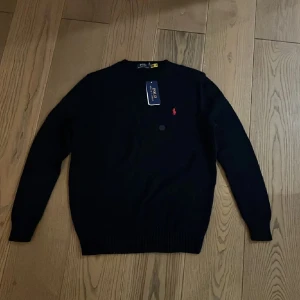 Svart tröja från Polo Ralph Lauren - Snygg svart tröja från Polo Ralph Lauren med det ikoniska röda logotypbroderiet på bröstet. Tröjan har långa ärmar och en klassisk rund halsringning. Perfekt för en stilren look.