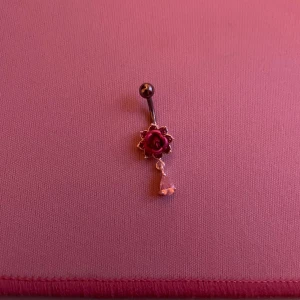 Navelpiercing med röd ros och kristall - Elegant navelpiercing med en röd ros i mitten och en hängande klar kristall. Staven är i silverfärg och har en rund kula på toppen. Perfekt för att ge en romantisk touch.