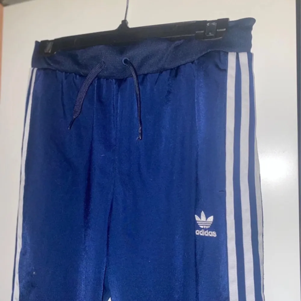Snygga blå träningsbyxor från Adidas med klassiska vita ränder längs sidorna. Byxorna har en bekväm resårmidja med snörning och broderade blommor på ena benet för en unik touch.. Farkut & Housut.