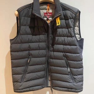 Svart dunväst från Parajumpers - Snygg svart dunväst från Parajumpers med dragkedja och hög krage. Västen har praktiska fickor med dragkedja och en gul detalj vid halsen. Perfekt för kyliga dagar.