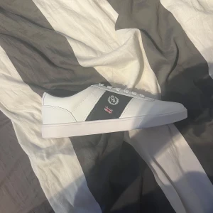 Vita sneakers från Henri Lloyd - Snygga vita sneakers från Henri Lloyd med svarta detaljer och snörning. Perfekta för en stilren look. Skorna har en diskret logga på sidan och en bekväm passform. Pris kan diskuteras