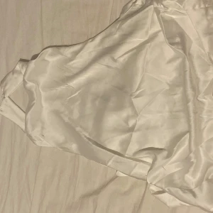 Vit axelbandslös topp - Elegant vit axelbandslös topp i ett mjukt och glansigt material. Perfekt för sommarens festligheter med sin stilrena design och knytning i midjan.