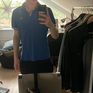 Blå pikétröja från Hugo Boss - Säljer en snygg blå pikétröja från Hugo Boss med modern passform. Tröjan har en klassisk krage med vita och svarta detaljer samt knappar framtill. Perfekt för en stilren look.