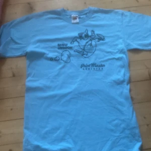 Blå t-shirt från Fruit of the Loom - Säljer en blå t-shirt från Fruit of the Loom med tryck på både fram- och baksidan. Framsidan har en tecknad figur och texten 'SISU UNGDOM'. Baksidan har texten 'KUL Å FLÅS I SISU FÅS!' samt flera logotyper och webbadress. Perfekt för en avslappnad stil.