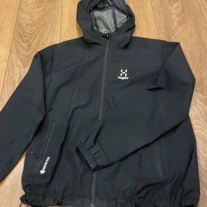 Svart vindjacka från Haglöfs - Säljer en svart vindjacka från Haglöfs i Gore-Tex-material. Jackan har en huva och dragkedja framtill samt två sidofickor med dragkedjor. Perfekt för utomhusaktiviteter med sitt slitstarka och vattentäta material. Nypris 2500!