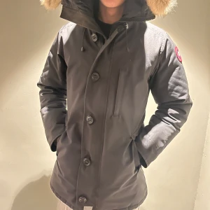 Svart parkajacka med pälsdetaljer - Säljer en stilren svart parkajacka från Canada Goose med pälsdetaljer på huvan. Jackan har knappar och dragkedja framtill samt en broderad logotyp på ärmen. Perfekt för kalla vinterdagar. Kvitto finns! Pälsen går att ta av om man hellre vill ha utan.