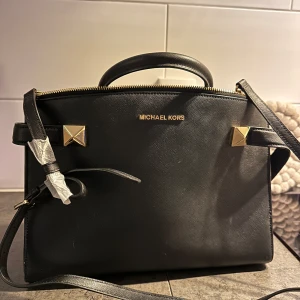 Svart handväska från Michael Kors - Elegant svart handväska från Michael Kors i skinn med guldfärgade detaljer. Väskan har en stilren design med två handtag och en avtagbar axelrem. Sparsamt använd. Några fläckar , går säkert få bort, jag har inte försökt. Mellanstorlek, en liten laptop får plats. Många fack.