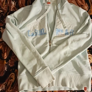 Ljusblå hoodie från Esprit - Säljer en ljusblå hoodie från Esprit med dragkedja och tryck i vitt och blått på framsidan. Tröjan har långa ärmar och ribbade muddar vid ärmslut och nederkant. Perfekt för en avslappnad stil!