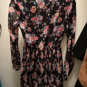 Blommig klänning med knytband - Snygg blommig klänning i svart med färgglada blommor. Den har långa ärmar och ett knytband vid halsen för extra stil. Perfekt för en chic look.