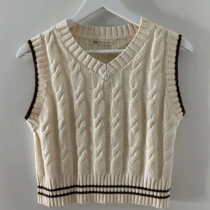 Beige stickad väst från STR - Säljer en snygg beige stickad väst från STR med kabelstickat mönster och bruna detaljer vid ärmhålen. Västen har en v-ringad design och ribbade kanter för en stilren look.✨✨