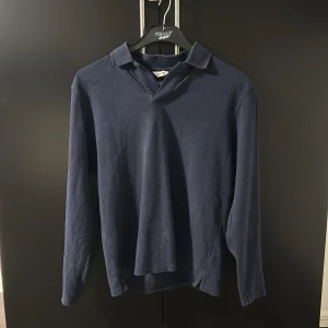 Mörkblå pikétröja från East West - Säljer en stilren mörkblå pikétröja från East West. Tröjan har långa ärmar och en klassisk krage. Perfekt för en avslappnad men ändå snygg look.