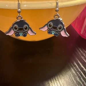 Söta örhängen med Stitch - Charmiga örhängen med en tecknad figur i svart och rosa. Perfekta för att ge en lekfull touch till din stil. Figurerna har stora ögon och utsträckta armar, vilket ger dem ett unikt och roligt utseende.