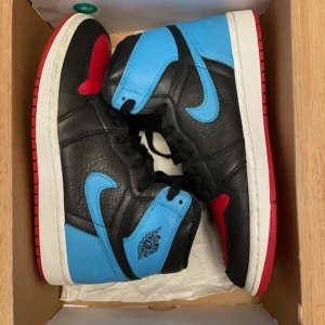 Nike Air Jordan 1 High OG UNC to Chicago - Snygga Nike Air Jordan 1 High OG i svart, blå och röd färgkombination. Skorna har en klassisk design med snörning och en hög siluett. Perfekta för sneaker-entusiaster som vill ha en ikonisk stil. Kommer med orginallåda