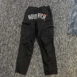 Svarta cargobyxor från Hoodrich - Snygga svarta cargobyxor från Hoodrich med elastisk midja och flera praktiska fickor. Perfekta för en avslappnad stil med en touch av streetwear. Byxorna har en cool logga på baksidan och är tillverkade i ett slitstarkt material.