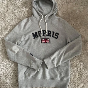 Morris hoodie - Snygg grå hoodie från Morris med broderad Union Jack-flagga och text på bröstet. Tröjan har en klassisk känguruficka och justerbar huva med snören. Säljer den pågrund av att jag har växt ur den. Storlek xs med en slimfitt. Hör av dig vid frågor eller funderingar!