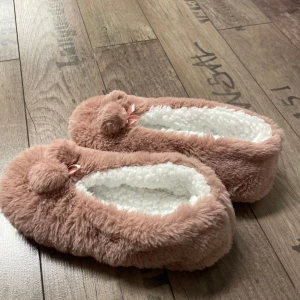 Bruna/rosa fluffiga tofflor med pom-poms - Mysiga bruna/rosa tofflor i fluffigt material med vita insidor. De har dekorativa pom-poms och små rosetter på ovansidan. Perfekta för att hålla fötterna varma och bekväma hemma.