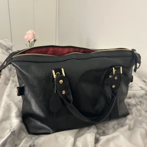 Svart handväska med dragkedja - Snygg svart handväska i skinn med guldfärgade detaljer och dragkedja. Väskan har ett rymligt rött foder och praktiska handtag. Den är riktigt fin och blir unik med röd insida. Får plats med mycket men enkelt att bära. 