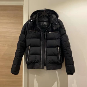 Svart dunjacka med huva - Snygg svart dunjacka från Moose Knuckles med avtagbar huva och dragkedja. Jackan har flera praktiska fickor med dragkedjor och en stilren design. Perfekt för kyliga dagar. Det finns 2 små snitt inuti luvan som inte syns och är lätt att laga. 