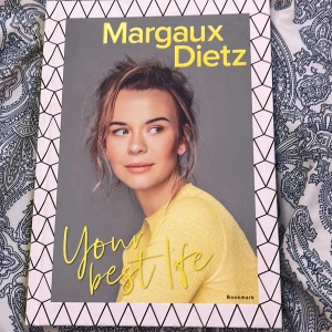 Your Best Life - Boken 'Your Best Life' av Margaux Dietz, utgiven av Bookmark, har en stilren design med ett porträtt av författaren på omslaget. Titeln är skriven i gult mot en grå bakgrund, vilket ger ett modernt och inbjudande intryck. Används intr