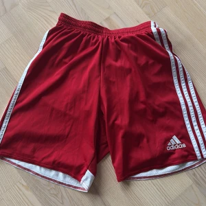 Röda shorts från Adidas - Snygga röda Adidas shorts med vita ränder längs sidorna och elastisk midja. Perfekta för sport och träning. Klassisk Adidas-logga på benet.