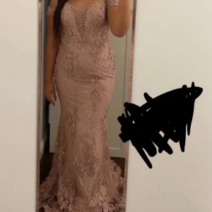 Rosa långklänning med spetsdetaljer - Elegant rosa långklänning med spets och pärlor. Klänningen har en v-ringad design och offshoulder-ärmar, vilket ger en sofistikerad look. Perfekt för festliga tillfällen.