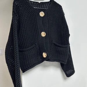 Svart stickad kofta med stora knappar - Snygg svart stickad kofta med stora guldknappar framtill. Koftan har en grovstickad struktur och två praktiska fickor. Perfekt för att hålla sig varm och stilren. Passar perfekt för s/m/l storlek.. Helt ny!