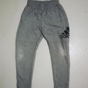 Grå mjukisbyxor från Adidas - Säljer ett par grå mjukisbyxor från Adidas med elastisk midja och svart logga på benet. Perfekta för en avslappnad stil eller träning. Byxorna har en bekväm passform och är tillverkade i mjukt material.