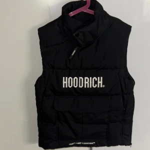 Svart dunväst från Hoodrich - Snygg svart dunväst från Hoodrich med dragkedja och tryckt logga på framsidan. Perfekt för att hålla värmen med stil. Västen har en hög krage och praktiska fickor.