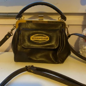 Mörkbrun handväska från Daniel Ray - Säljer cool mörkbrun handväska i skinn från Daniel Ray med guldfärgade detaljer. Väskan har en praktisk dragkedja. Väskan kommer inte till användningdärför säljer ja den. Helt ny har aldrig använts 