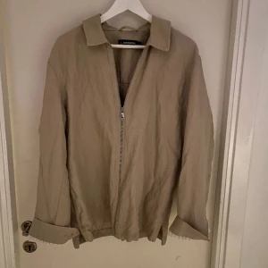 Beige jacka från J.Lindeberg - Snygg beige jacka från J.Lindeberg med dragkedja framtill. Jackan har en klassisk krage och långa ärmar med uppvikta ärmslut. Perfekt för en stilren look.