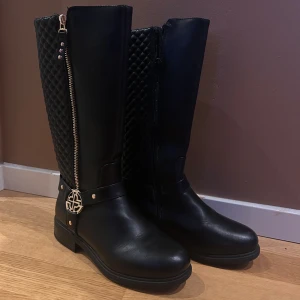 Svarta stövlar med guld-dragkedja i storlek 37 - Snygga svarta stövlar i skinn med quiltad baksida och dekorativ dragkedja. De har en elegant metallspänne-detalj och är perfekta för höst och vinter. Bara använda 1 gång. 