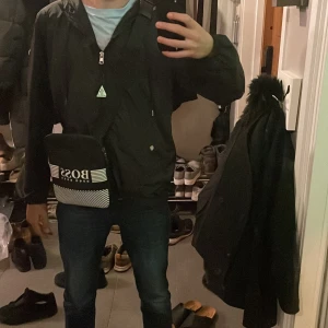 Svart axelväska från Hugo Boss - Snygg svart axelväska från Hugo Boss med vit logotyp och randigt mönster. Perfekt för att bära dina nödvändigheter med stil. Väskan har en praktisk dragkedja för säker förvaring.