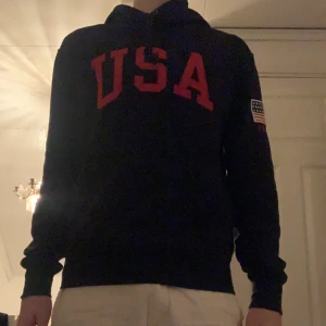Ralph Lauren mörkblå hoodie med USA-tryck - Snygg mörkblå Ralph lauren hoodie med rött USA-tryck på bröstet och amerikansk flagga på ärmen. Perfekt för en avslappnad stil.