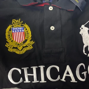 Svart pikétröja från Ralph Lauren - Snygg svart pikétröja från Ralph Lauren med stora broderade detaljer. Tröjan har texten 'CHICAGO' i vitt och ett emblem på bröstet. Klassisk krage med knappar och en broderad polospelare i vitt. Chief Keef Polo.