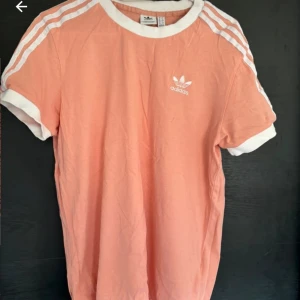 Persikofärgad t-shirt från Adidas - Snygg persikofärgad t-shirt från Adidas med vita detaljer och klassiska tre ränder på axlarna. Den har korta ärmar och en rund halsringning. Perfekt för en sportig look!