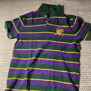 Randig pikétröja från Ralph Lauren - Säljer en snygg randig pikétröja från Ralph Lauren i grönt, lila och gult. Tröjan har korta ärmar och en klassisk krage med knappar. Broderad logga på bröstet för en stilren look. Perfekt för en avslappnad stil!