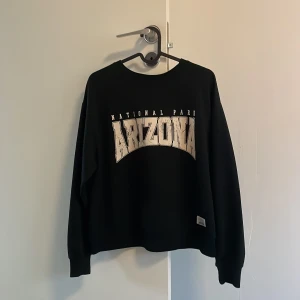 Svart tröja med Arizona-tryck från DF Young - Säljer en svart tröja från DF Young med stort Arizona-tryck på framsidan. Tröjan har långa ärmar och en klassisk rund halsringning. Perfekt för en avslappnad stil.Det är inga problem på den och ser ut som ny.