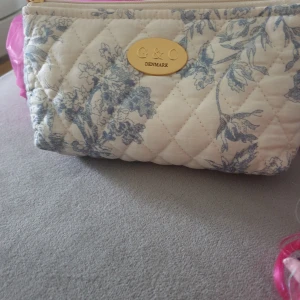 Quiltad blommig väska från G&C Denmark - Snygg quiltad väska med blommigt mönster i blått och vitt från G&C Denmark. Väskan har en dragkedja upptill och en elegant guldplatta med logotyp. Perfekt för att hålla dina småsaker organiserade.