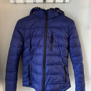 Blå pufferjacka från H Collection  - Snygg blå pufferjacka från H Collection med dragkedja och huva. Perfekt för kyliga dagar med sin vadderade design och praktiska fickor med dragkedjor. En stilren jacka som håller dig varm och bekväm.