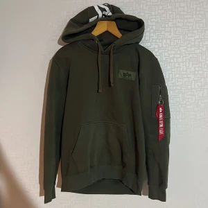 Grön hoodie från Alpha Industries - Säljer en grön hoodie från Alpha Industries med stor logga på ryggen och en mindre på bröstet. Hoodien har en praktisk ficka med dragkedja på ärmen och justerbar huva med snören. Perfekt för en avslappnad stil.
