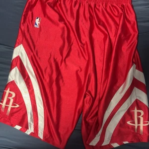 Röda basketshorts med NBA-logga - Snygga röda basketshorts med vit randig design och NBA-logga. Perfekta för sport och träning. Elastisk midja för bekväm passform.