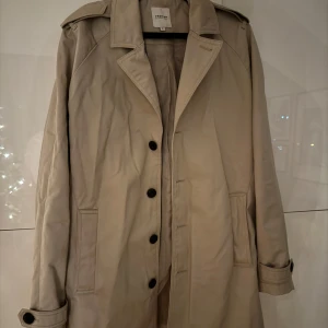 Beige trenchcoat från Jack & Jones - Snygg beige trenchcoat från Jack & Jones i storlek M. Jackan har klassiska knappar framtill och en stilren design med långa ärmar. Perfekt för en elegant look.