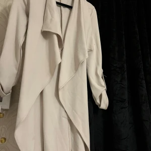 Beige trenchcoat med knytband - Elegant beige trenchcoat med långa ärmar och ett stilrent knytband i midjan. Jackan har en flytande design framtill och är perfekt för vår och höst. En tidlös klassiker för garderoben.