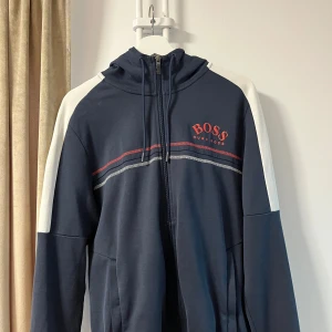 Mörkblå huvtröja från Hugo Boss - Snygg mörkblå huvtröja från Hugo Boss med vita ärmar och röd logga på bröstet. Tröjan har dragkedja och en huva med röda detaljer. Perfekt för en avslappnad stil.