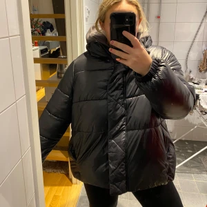 Svart pufferjacka med huva - Säljer en svart pufferjacka med huva. Jackan har en glansig yta och dragkedja framtill. Perfekt för kalla dagar med sin vadderade design. Storlek XS men passar upp till L.