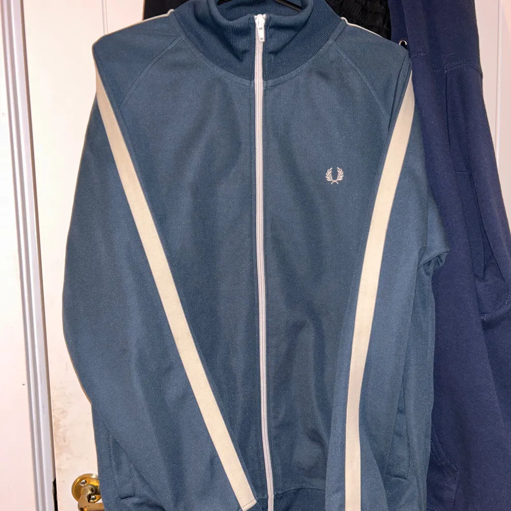 Snygg blå track jacket från Fred Perry med vita detaljer och dragkedja. Perfekt för en sportig look eller avslappnad stil. Jackan har långa ärmar och en klassisk krage.. Takit.