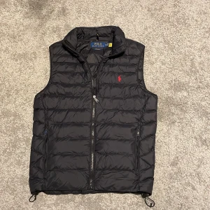 Svart dunväst från Polo Ralph Lauren - Säljer en snygg svart dunväst från Polo Ralph Lauren. Västen har en dragkedja framtill och är dekorerad med det klassiska röda Polo-logotypen på bröstet. Perfekt för lager-på-lager under höst och vår. Kan diskutera pris. Skick 8,5/10. Kan ta byten.